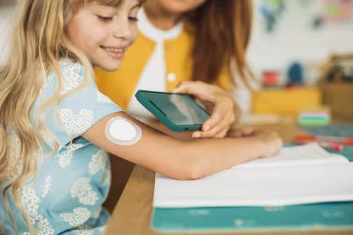 Eine Diabetesbegleitung misst den Blutzucker am Armsensor eines Mädchens.